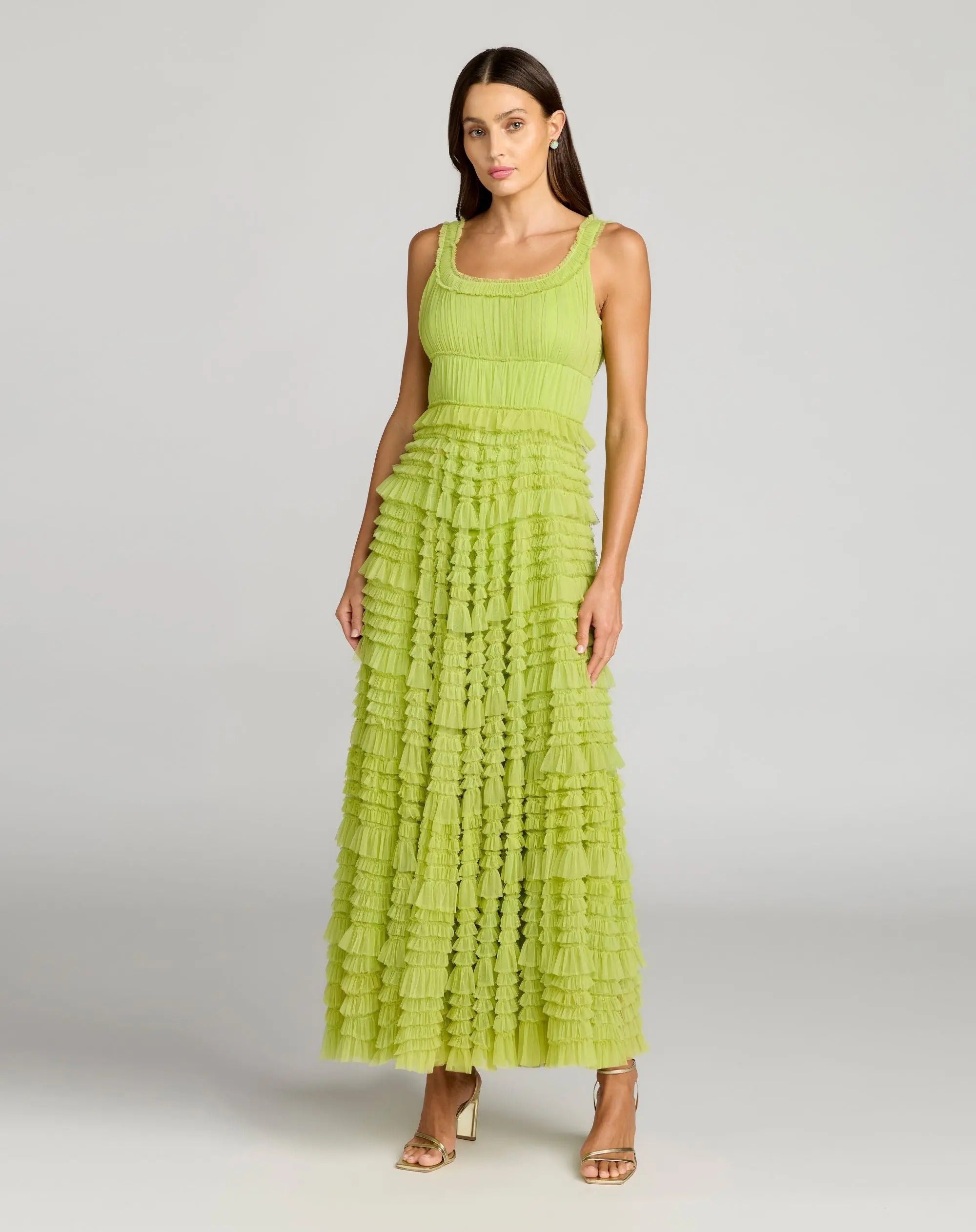 A woman stands against a plain light gray background wearing the Mac Duggal 8292 Tea-Length Party Dress—a sleeveless, lime green A-line tiered maxi with delicate ruffle detailing—paired with gold open-toe heels.